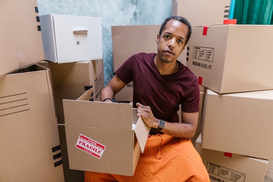 A young man with short curly hair, wearing a maroon t-shirt and bright orange trousers, stands amid a collection of cardboard boxes in an indoor storage or packing area. He is holding and looking at a