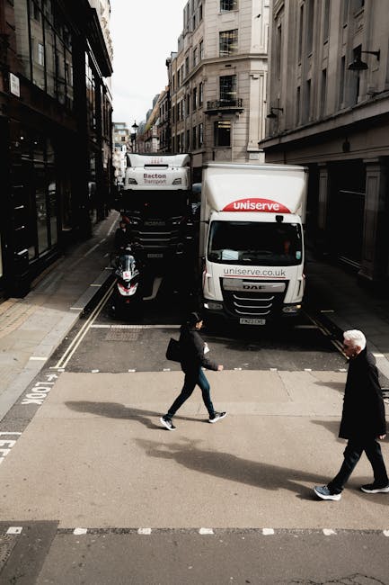 A street scene showing parked white vans and cars along the pavement near a white building with decorative architectural details. The pavement is lined with black and white bollards, and fallen autumn