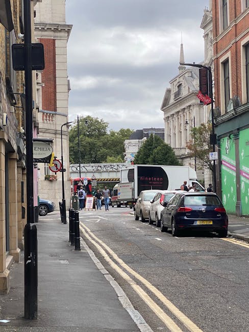 A narrow street in a residential area with parked cars along the curb, some with visible registration plates, and a white moving van parked near the pavement. Several cardboard boxes and wrapped furni