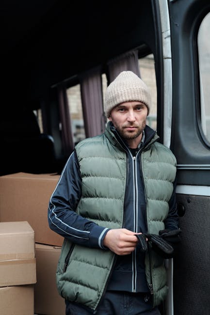 A man wearing a dark vest, black trousers, and a cap is loading cardboard boxes into the back of a large white moving van parked outside a commercial building. The van's side doors are open, revealing