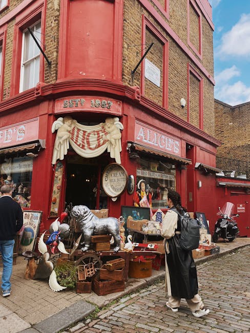The exterior of a historic red-brick building with a rounded corner features signage for a shop named 'Alices,' with decorative white cherub sculptures and a banner above the entrance. Outside, there are various vintage items and antiques displayed on wooden crates and tables, including a large plush dog and ceramic geese, with some objects partially visible on the sidewalk. A woman dressed in dark clothing and a backpack stands nearby, observing the display. The building's windows and door are visible, with black awnings above the shop entrances, and the cobbled street and pavement surround the scene. Bright daylight illuminates the scene, suggesting daytime urban shopping or browsing, and the overall setting hints at a lively, historic shopping street typical of London or Ladbroke Grove areas, supporting house removal and packing processes when relocating nearby.