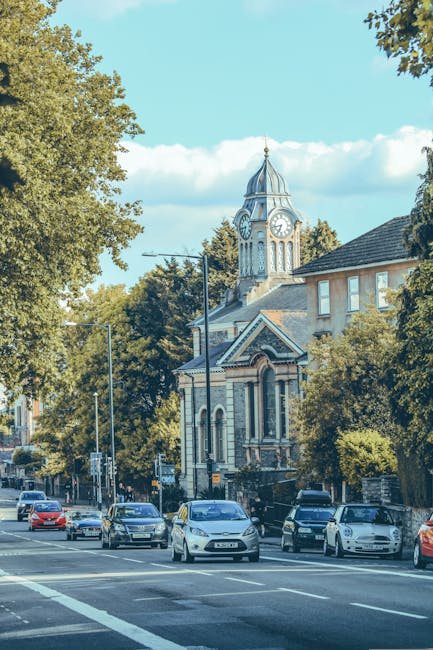 A street scene in Ladbroke Grove showing several parked cars along the pavement, with a historic church featuring a clock tower and a pointed spire in the background, surrounded by lush green trees. The church's architecture includes arched windows and decorative brickwork, set against a clear blue sky. The road is busy with moving vehicles, and the area appears to be part of a residential and commercial neighbourhood. Man and Van Ladbroke Grove provides professional house removals and furniture transport services, supporting clients with packing, loading, and moving logistics during home relocations within the area.