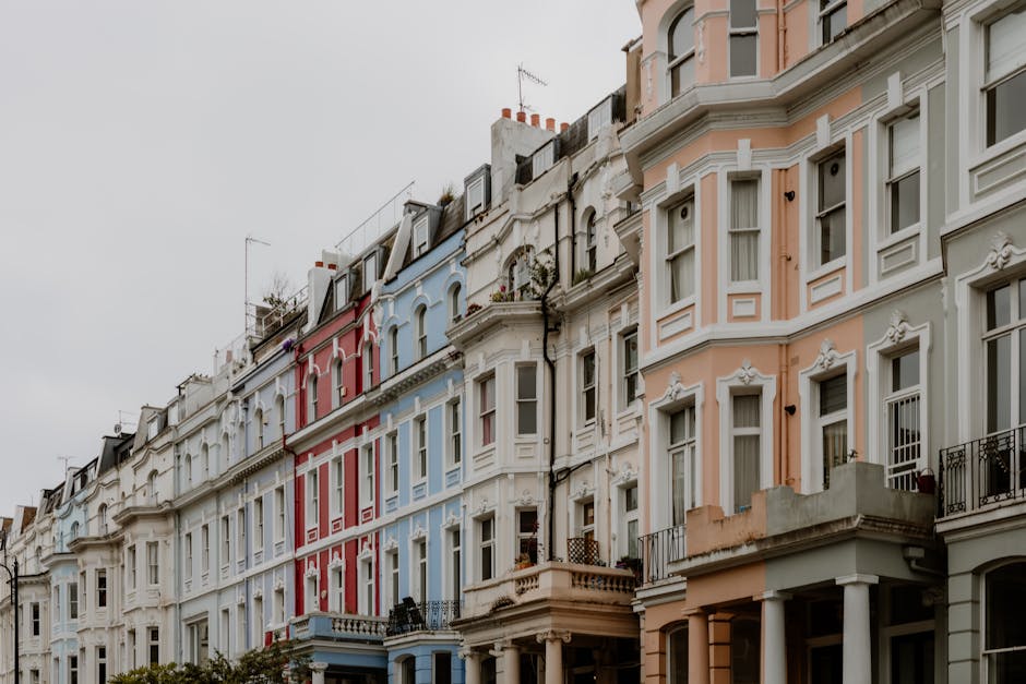 Photograph of a row of Victorian terraced houses along Westbourne Grove, showing multiple stories with large sash windows and decorative stucco facades painted in pastel shades of pink, white, blue, and cream. The buildings have small balconies with black wrought iron railings, some with potted plants or outdoor furniture. The street scene captures the outdoor environment with overcast sky, and the facades are detailed with ornate cornices and moldings. The image is taken from street level, focusing on the uniformity and architectural style of the residential buildings, suggesting an area where home relocation and furniture transport are common. The scene conveys a typical London residential street suitable for visual context about house removals and packing and moving services, as provided by Man and Van Ladbroke Grove, designed to assist W10 residents with their moving needs. The overall setting emphasizes the urban environment and the typical exterior appearance of homes involved in removals and transport tasks.