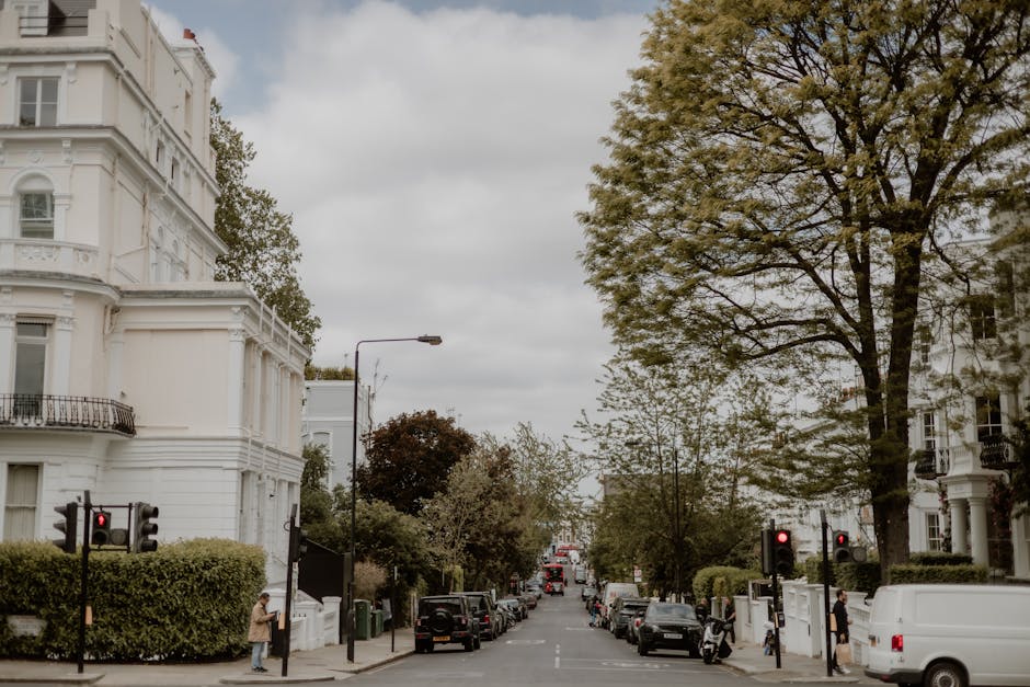 Image of a residential street on a clear day, featuring a row of elegant, multi-storey Georgian-style buildings with cream and beige facades, decorative window frames, and wrought-iron balconies. Several white and brown cardboard boxes, some wrapped in clear plastic or brown paper, are placed on the pavement near the buildings, suggesting packing materials for a home relocation. In front of the buildings, a small delivery trolley is visible, loaded with additional boxes and moving supplies, indicating active packing or loading activities. Parked black and dark-colored cars line the street, while a black van with an open rear door is positioned on the curbside, part of the moving process by Man and Van Ladbroke Grove. The van appears to be in the midst of loading or unloading furniture, boxes, or packing materials, with a few workers possibly nearby, although not clearly visible. The environment is well-lit with natural daylight, and the scene captures the typical setting for furniture transport or house removals in Westbourne Grove, W10, highlighting the logistics involved in home relocation services.